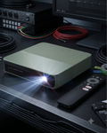 A compact rectangular projector in a muted sage green color sits on a dark desk, casting a bright beam of light forward. A black remote control rests beside it, with cables, audio equipment, and a monitor in the background.